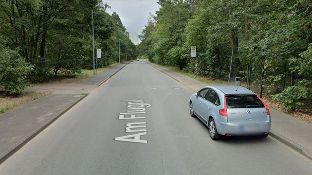 Google-StreetView-Bild. Straße Am Flugplatz, etwa zweistreifig, 2-m-Bürgersteige links und rechts. In mittlerer Entfernung Bushaltestellen, direkt davor zu beiden Seietn abgesenkte Bordsteine, wo ein Waldweg kreuzt