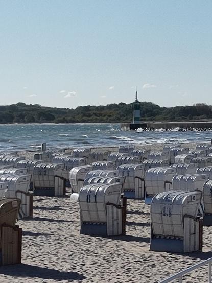 Weiße Strandkörbe, sonniges Meer mit Schaumkronen, grün weißer Leuchtturm.
