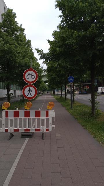 Ein Sperrelement steht auf einem Fuß-/Radweg. Darüber Schilder für Radfahrer und Fußgänger gesperrt. Dahinter sichtbar ein Schild benutzungspflichtiger Radweg.