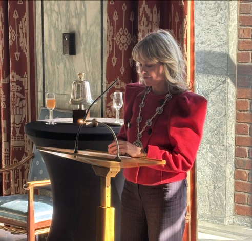 Oslo mayor Anne Lindboe speaking at a lectern at the City Hall at thr EASE welcome reception. She is a white woman with shoulder length blonde hair, wearing a bright red jacket with shoulder pads, a gold mayoral chain, and grey trousers.