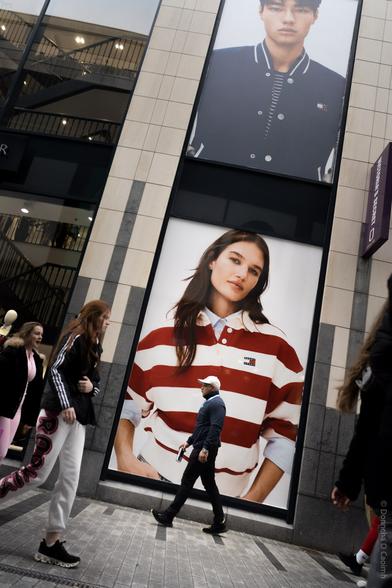 A candid street photograph taken in Cork City, Ireland, showing people walking past a large retail storefront with two oversized fashion posters. One poster features a person in a navy varsity jacket, and the other shows a person in a red and white striped rugby shirt, both displaying the Tommy Hilfiger logo. The scene captures urban life and the intersection of fashion advertising and everyday movement.