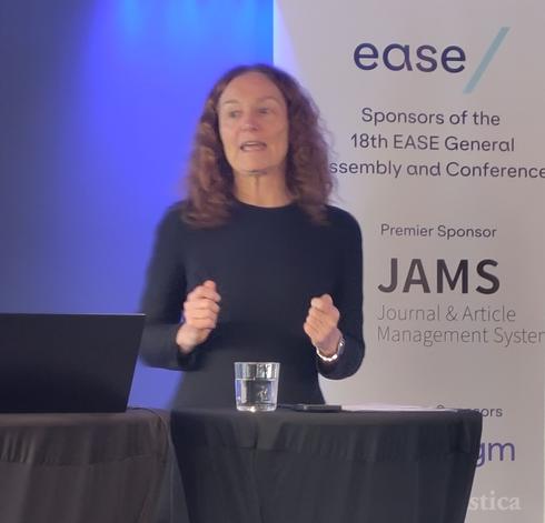 Camilla Stoltenberg speaking at the EASE conference podium. She is a white woman with shoulder length curly brown hair, wearing a long sleeved black top.