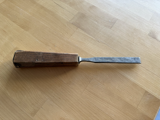 A chisel on a table. The handle is blocky and square-ish, and beaten up at the hitting end. The iron is a shiny grey