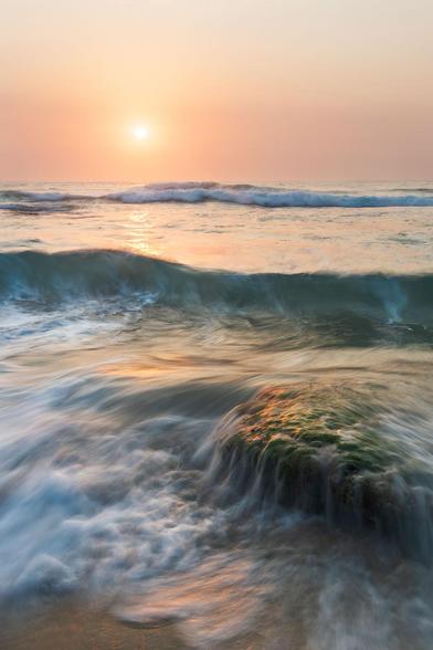 a vertical sunrise seascape in peach colours and incoming standing wave. Another wave is flowing down the mossy rock in the foreground. The horizon is misty or smoky and the sun is visible as a diffused peach blob