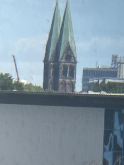 Image features a tall, historical building with two green spires, partially obscured by a modern structure in the foreground. The sky is clear and blue.