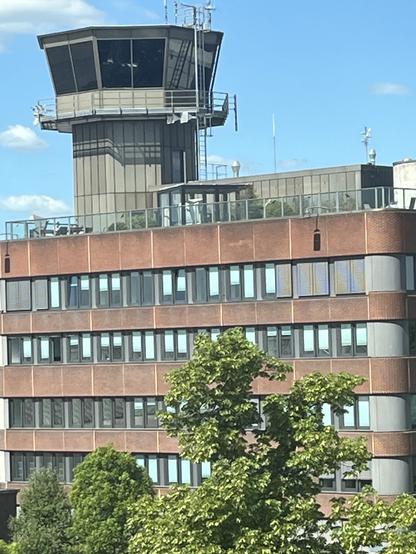 An airport control tower is visible atop a multi-story building, with a clear blue sky in the background and some trees in the foreground. The building features numerous windows and a rooftop terrace.