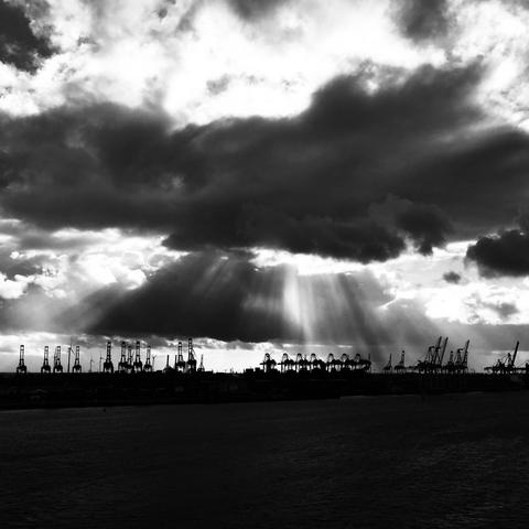 black and white photograph of the cranes (sea dogs) of hamburg harbour silhouetted against a dramatic sky. rays of light are seen through large black clouds. drama!