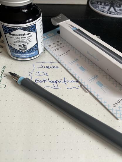 Una pluma de inmersión que no deja de ser un cuerpo de plástico sencillo con un plumín al final. La pluma es gris con algún pequeño detalle azul. Está apoyada sobre una libreta donde se ha escrito el texto “Jueves de estilográficas” en letras azules. Al fondo un bote de tinta de cristal y la caja de cartón de la pluma.