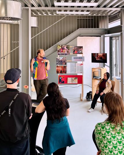 Ein Mann steht gestikulierend vor einer Anzeigetafel, während ein kleines Publikum zuhört. Der Schauplatz scheint ein moderner Innenraum mit einer Treppe im Hintergrund zu sein, und auf der Anzeigetafel sind verschiedene Informationsposter und Bilder zu sehen. Ein Zuhörerin sitzt auf einem Stuhl vor der Anzeigetafel.