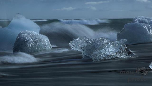 The image shows a dramatic scene of ice chunks scattered on a black sand beach with waves crashing in the background. The ice appears to be glistening, likely due to sunlight, creating a striking contrast with the dark sand and water. This type of scenery is often found in places like Iceland's Diamond Beach.