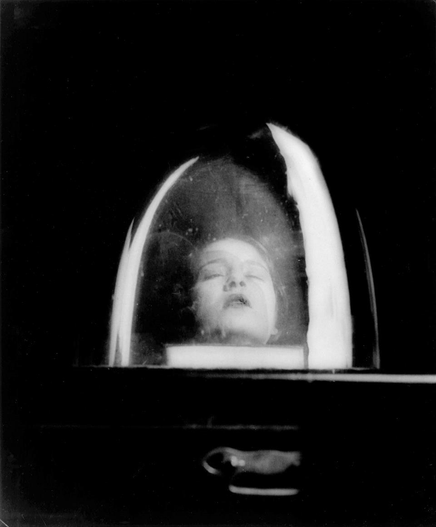 sw Foto
Auf einem Tisch steht eine Glaskuppel. Es sieht so aus, als ob darin der Kopf einer Frau liegt. Sie hat die Augen geschlossen.