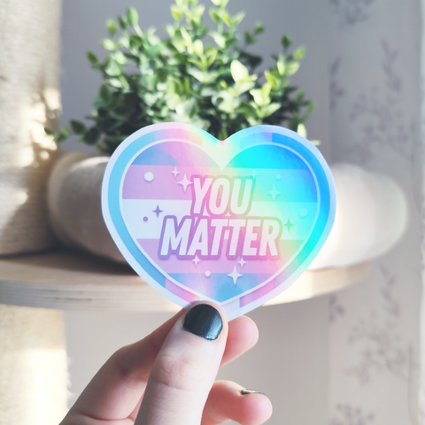 hand holding up a holographic sticker of a heart with trans flag colors and text "you matter"