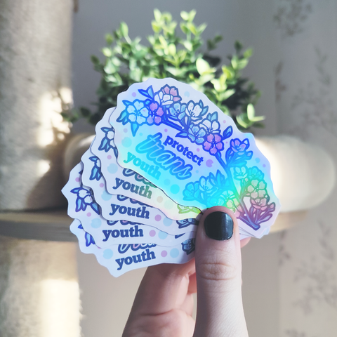 hand holding up a holographic sticker of a branch blooming in trans flag colors titled "protect trans youth"