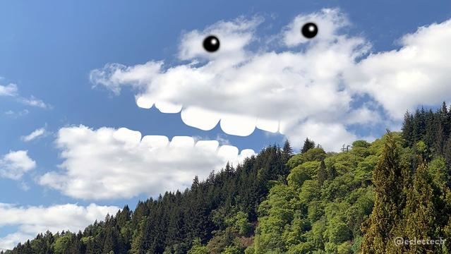 A photo of a wooded hillside sloping up on the right hand side. The trees are different shades of green, and bright in the sunlight. The sky above is blue, with some fluffy white clouds, some of which are very vaguely shaped like a head with an open jaw and two bulbous eyes on top. I have added teeth and eyes.
