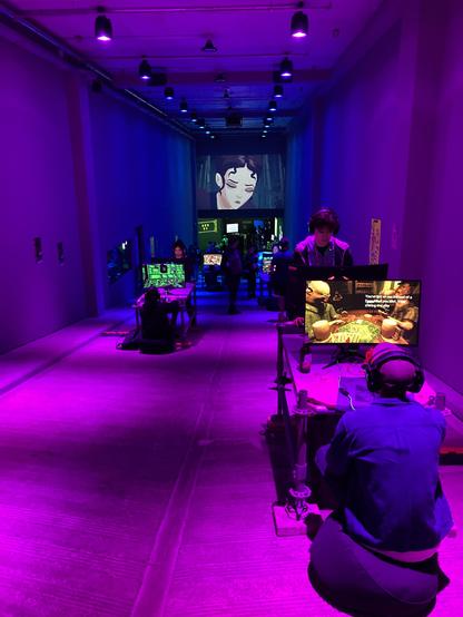 a photo of a purple and blue-lit indoor space. people are sitting and standing in front of video game demos at amaze festival 2025. theres a large screen above, showing the face of a video game avatar in close up.