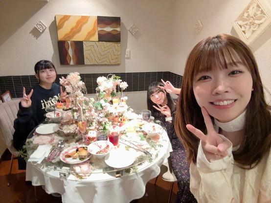 Three women are enjoying a festive meal together at a beautifully decorated table with a floral and dessert-themed setup. They are all smiling and making peace signs, creating a cheerful and friendly atmosphere. The room’s warm lighting and elegant decor enhance the intimate and celebratory feel of the gathering.