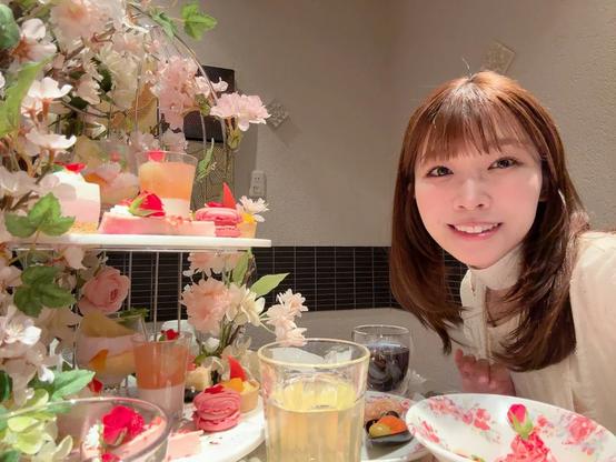 A woman smiles warmly at the camera during what appears to be a beautifully arranged dessert gathering. The table is adorned with floral decorations and an elegant display of pastel-colored treats including macarons, fruit tarts, and layered mousse. The overall ambiance is cozy and festive, perfect for a cheerful and intimate celebration.