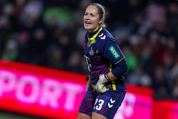 Aurora Mikalsen steht während des Spiels leicht vorn über gebeugt und ruft mit leicht gerunzelter Stirn etwas in die Ferne. Ihr Torhüterinnen Trikot ist violett und trägt das Emblem des 1. FC Köln, hinter ihr leuchtet unscharf eine Werbebande in pink und rot.