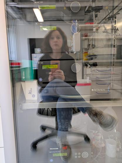 Me, a white dark haired woman, sitting on a chair in a lab and taking a picture of my reflection in the glass door of a cooling cabinet.