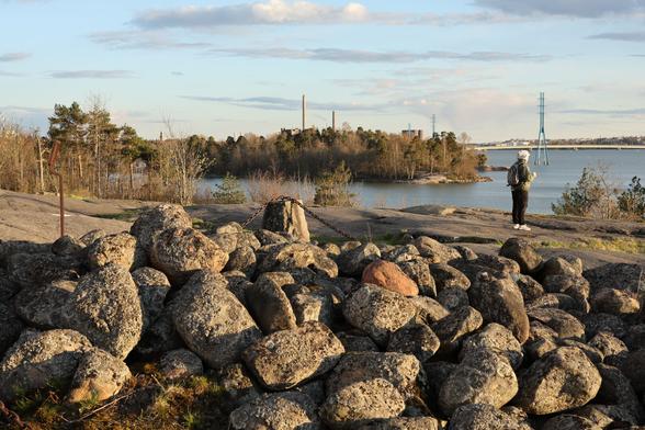 A photo of a Bronze Age cairn and the coastal cliffs in Meilahti on a sunny spring evening. There is a person standing on the right.
