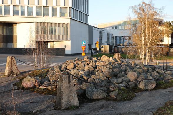 A photo of a Bronze Age cairn and an intersection and a hospital behind it.