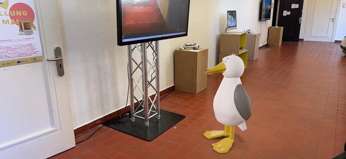 Eine AR Steinmöwe (Cartoonzeichnung) ist im Umfeld der Ebene 0 im Altbau der Muthesius Kunsthochschule beben einigen Monitoren in einem Flur dargestellt.