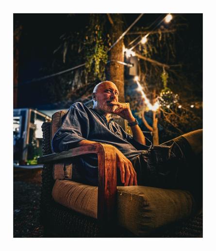 The photo is of a follicularly challenged, bearded man sitting in an armchair. He is sitting outdoors at night. There are string fairy lights visible in the background. The is wearing a blue shirt with the sleeves rolled up and some shorts. He is sitting parallel to the picture but is looking just above the centre of the image. He has one arm resting on the armrest and his left arm, (furthest from us) is bent with fingers covering his mouth.