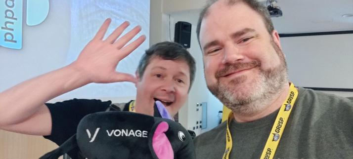 Me and James Seconde posing with a Vonage Elephpant (black plushie elephant with the inside of the ears colored, one purple, the other pink, Vonage and their logo printed in white on the visible side) with a PHPDay presentation slide in the background.