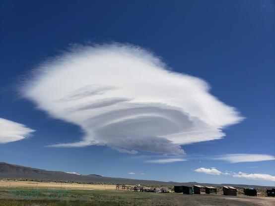 Four layer lenticular cloud, of which only the bottom two layers of round lens like cloud is clearly defined and the other two out layers can be seen by their left edges as the top part of the cloud mushrooms out in a fuzzy all, in a dark blue sky. A line of beaten trash dumpsters can be seen in the bottom right of the image. Photo by Jenifer Hanen.