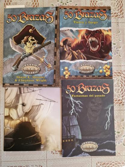 La imagen muestra cuatro libros de la línea "50 Brazas" de Savage Worlds. Cada libro tiene una portada con ilustraciones diferentes. El primero muestra una calavera pirata con pistolas cruzadas. El segundo muestra una criatura roja monstruosa y a un hombre con una espada. El tercero muestra un barco de vela antiguo en una escena brumosa. El cuarto muestra un barco en una tormenta con una figura espectral amenazante. Todos los libros incluyen el logotipo de Savage Worlds y los títulos en español, indicando que son diferentes aventuras de la misma serie.