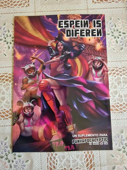 La portada de un suplemento para el juego de rol "Punkapocalyptic" muestra a una mujer con ropa de motociclista y armadura, en pose heroica, con una capa roja y negra ondeando detrás de ella. Tres criaturas antropomórficas con trajes blancos y cascos, que parecen ser acompañantes o aliados, la flanquean. Al fondo, se ve un paisaje apocalíptico con llamas y un edificio que parece una torre o monumento. El título "ESPEIN IS DIFEREN" está escrito en la parte superior en letras estilizadas. En la parte inferior, se lee "Un suplemento para Punkapocalyptic, el juego de rol".