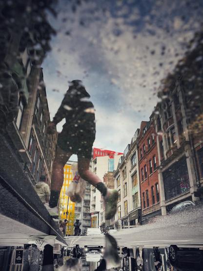 Eine städtische Straße spiegelt sich in einer Pfütze, wodurch die Perspektive auf den Kopf gestellt wird. In der Reflexion sieht man eine Person in dunkler Kleidung, die eine Tasche trägt und über die Straße geht. Die umgebenden Gebäude sind vielstöckig und haben unterschiedliche architektonische Stile, einige mit auffälligen roten und gelben Fassaden. Autos sind ebenfalls in der Reflexion zu erkennen.