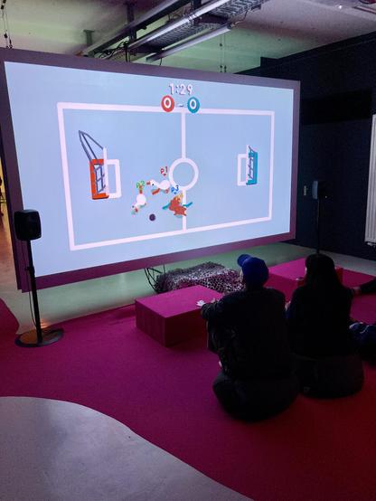 a photo of several people sitting on a red carpet in front of a large screen. thescreen shows a video game scene: a blue playing field withtwo opposing teams (geese and capybaras) playing cricket