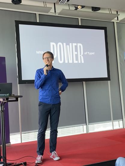 Oliver in a blue shirt on stage is in front of a large screen displaying the text "With the POWER of Type!" on a blue background.