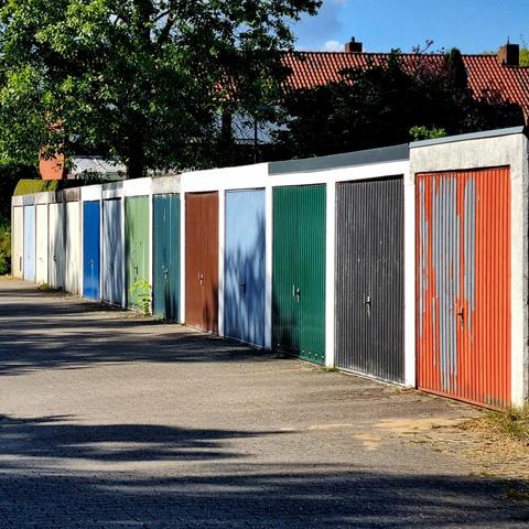 A dozen closed garage doors in a row, each in a different color. The shade of trees falls on some of them. Behind the garages are trees and houses with tiled roofs.