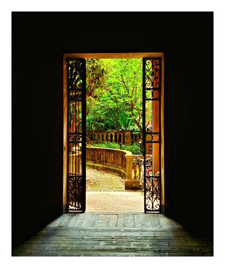 The photo is of a doorway with ornate metal gates on either side of the doorframe leading to a sunlit courtyard garden. The courtyard has a curved stone pathway, balustrades, greenery, and there is a bicycle parked on the right hand side. There is a big contrast between outside and inside. The outside is all sunlit but the inside is black and shadowy.