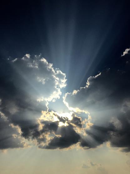 A sky scene featuring dramatic crepuscular rays. The sun, obscured behind a cluster of dark, billowing clouds, emits powerful beams of light that radiate outward in all directions. These sunbeams pierce through gaps in the cloud cover, creating a striking contrast between light and shadow. The clouds are a mix of deep grays and bright, illuminated edges where the sunlight catches them. The sky transitions from a deep, moody blue at the top to softer, warmer hues near the horizon.