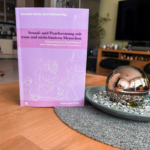 Das Buch “Sexual- und Paarberatung mit trans und nicht-binären Menschen. Praxisorientierte Empfehlungen und erfahrungsbasierte Perspektiven” steht auf meinen Wohnzimmertisch neben einer Silberkugel, die sich in einer Schale mit grauem und grünen Kies befindet. Das Cover zeigt weiß stilisierte Menschen bei diversen sexuellen Praktiken auf einem violetten Hintergrund.