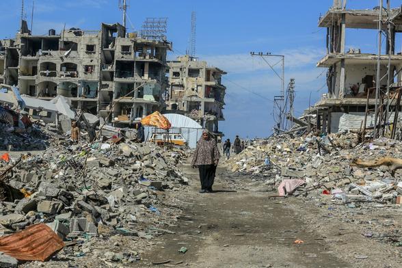 A woman in a patterned scarf walks through a devastated landscape filled with rubble and debris from destroyed buildings. Several individuals can be seen in the background amidst the destruction, under a blue sky.

Rafah February 2025