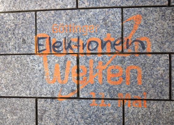 Stencil-style graffito on a sidewalk, advertising a quantum physics event called 'Goettinger Quantenwelten'. Someone wrote the word 'Elektronen' (electrons) over the word Quanten with a black marker.