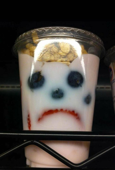 A yogurt parfait cup with granola on top appears to have a sad face formed by blueberries and a curved slice of strawberry.