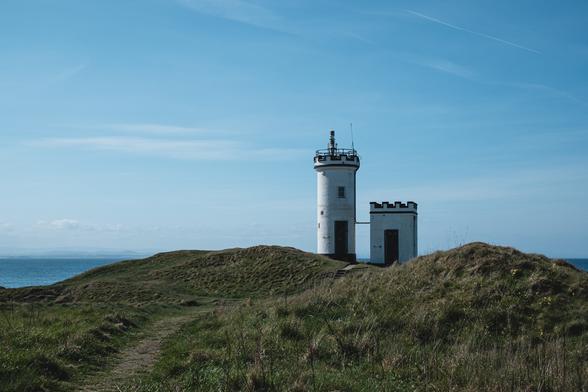 A lighthouse stands on a small green promontory. The lighthouse consists of one tall vertical tower and one smaller oblong tower. The sky is blue.