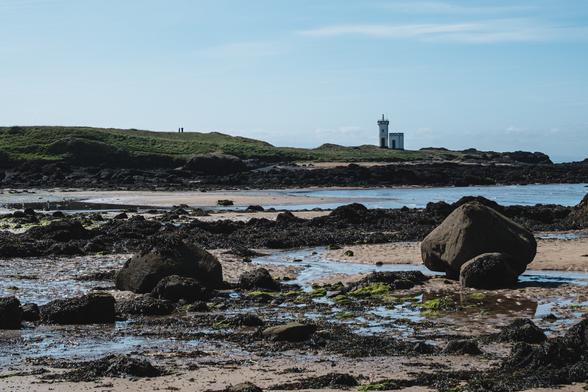 My photograph of a beach with the tide out there are many large boulders across the beach and in the distance is a lighthouse. The sky is blue.