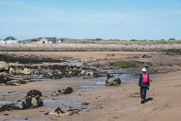A photograph of a beach with large boulders and a seawall in the distance. There is a single figure walking across the beach with a red rucksack and a white hat.