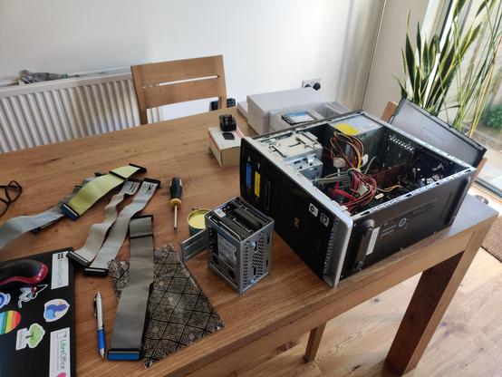 A wide shot of a kitchen table with a desktop computer opened up and lying on its side. The hard drive cage contains an IDE and a SATA hard disk. IDE and floppy ribbon cables are strewn on the table.