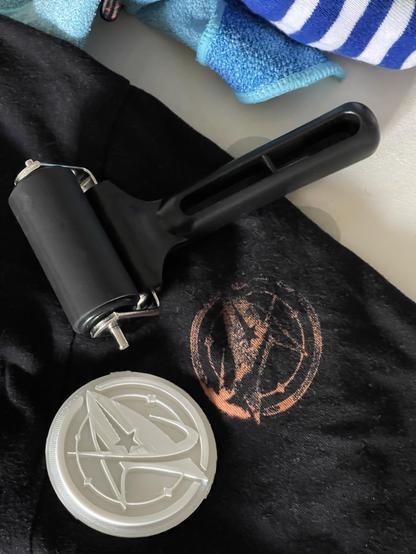 An image showing a black t-shirt with an orange printed design, a printing roller, and a circular stamp featuring a logo with a star and circular outline. Blue and white striped fabric and a blue washcloth are in the background.