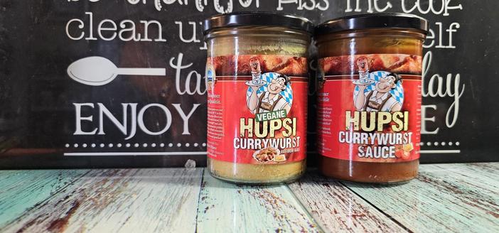 Das Bild zeigt zwei Glasflaschen mit Hupsi Currywurst-Sauce, die auf einem Holztisch stehen. Die Flaschen haben rote Etiketten mit dem Logo eines Mannes in Tracht, der eine Currywurst hält. Auf der linken Flasche steht "Hupsi Currywurst" und auf der rechten "Hupsi Currywurst Sauce". Die Flaschen sind mit schwarzen Korken verschlossen. Im Hintergrund ist ein schwarzes Brett mit weißen Schriftzügen zu sehen, die "clean up, taste, enjoy" und "tasty day" enthalten, sowie ein Zeichen eines Löffels.