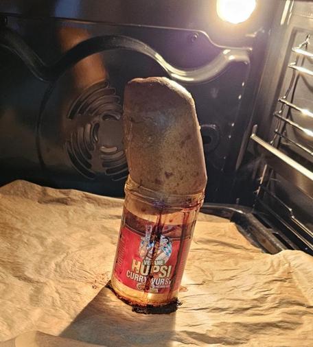 Das Bild zeigt eine Currywurst, die in einem offenen Ofen auf einem Backpapier steht. Die Currywurst ist auf einem durchsichtige Glas mit der Aufschrift "VEGANE HUPSI CURRYWURST" gesteckt, die auf dem Boden des Ofens liegt. Die Flasche enthält vermutlich Currywurst, da die Flüssigkeit aus der Flasche auf das Backpapier tropft. Der Ofen hat eine Metallwand mit einem Ventilator und ein Licht, das das Innere des Ofens beleuchtet. Die Currywurst ist mit der Oberseite nach oben in der Flasche stecken geblieben, wodurch sie wie ein Fels in der Flut wirkt.
