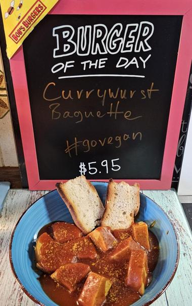 Das Bild zeigt ein Gericht namens "Currywurst Baguette" als "Burger of the Day" bei Bob's Burgers. Das Gericht besteht aus einer Baguette, die in Stücke geschnitten und in einer Currywurst-Sauce mit Gewürzen angerichtet ist. Die Currywurst-Sauce ist braun und glänzend, und die Baguettestücke sind in der Sauce getaucht. Neben dem Gericht befindet sich ein schwarzes Tafel-Schild mit rotem Rahmen, auf dem in weißer und gelber Schrift "BURGER OF THE DAY" und "Currywurst Baguette #govegan $5.95" steht. Das Schild ist mit einem gelben Sticker mit der Aufschrift "BOB'S BURGERS" verziert. Das Gericht ist in einem blauen Teller serviert, der auf einem Holztisch steht.
