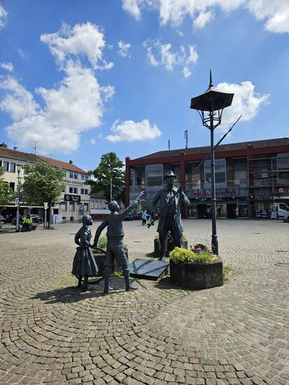 Das Denkmal auf dem Marktplatz Dudweiler. Der Mann, der früher die Gaslampen anzündete. Daneben zwei Kinder die ihm zuschauen. Dahinter sieht man einen blauen Himmel mit wenigen weißen Wolken.